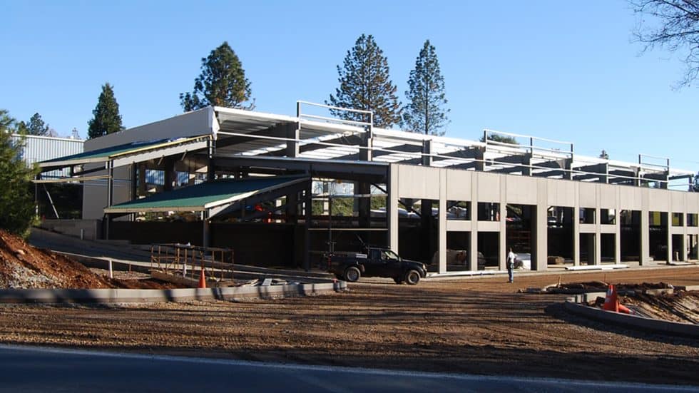 Modern multi-story commercial building under construction, showcasing sleek architectural design and structural framework, with a scenic backdrop of tall trees and a clear blue sky.