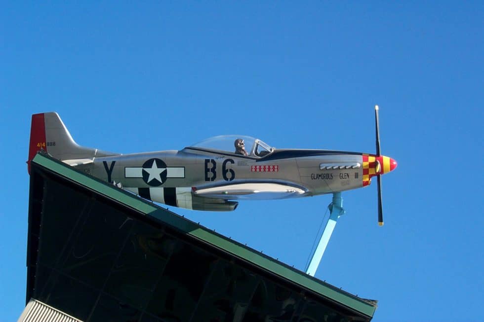 Fighter jet airplane mounted on a rooftop display against bright blue sky, featuring vintage military markings, colorful nose art, and a pilot figure in the cockpit.